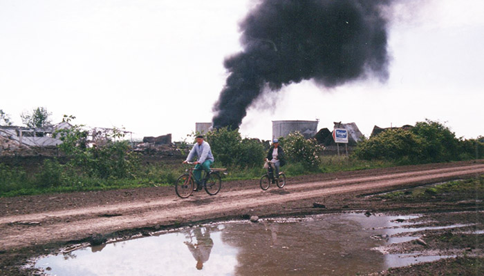 Two people are riding bikes, and behind them you can see the consequences of NATO bombing of Serbia.